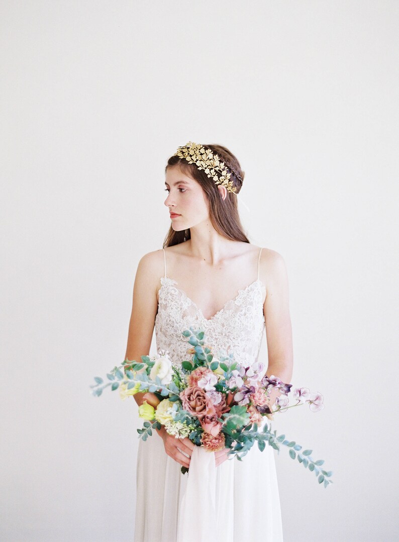May include: A bride wearing a white lace dress and a gold leaf crown holds a bouquet of pink, purple, and yellow flowers.