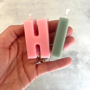以下が含まれることがあります： 「HI」の文字を形どった2つのキャンドル。 「H」のキャンドルはピンク色で、「I」はくすんだ緑色です。どちらのキャンドルにも芯があります。キャンドルは手で持たれています。