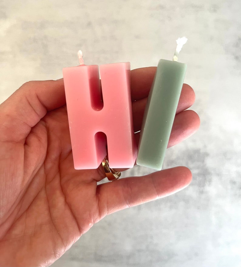 以下が含まれることがあります： 「HI」の文字を形どった2つのキャンドル。 「H」のキャンドルはピンク色で、「I」はくすんだ緑色です。どちらのキャンドルにも芯があります。キャンドルは手で持たれています。