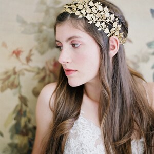 May include: A woman with long brown hair wears a gold floral crown headband. The crown is made of delicate metal leaves and flowers. She is wearing a white lace top.