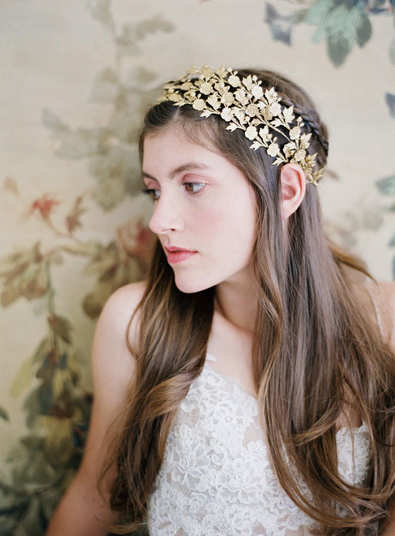 May include: A woman with long brown hair wears a gold floral crown headband. The crown is made of delicate metal leaves and flowers. She is wearing a white lace top.