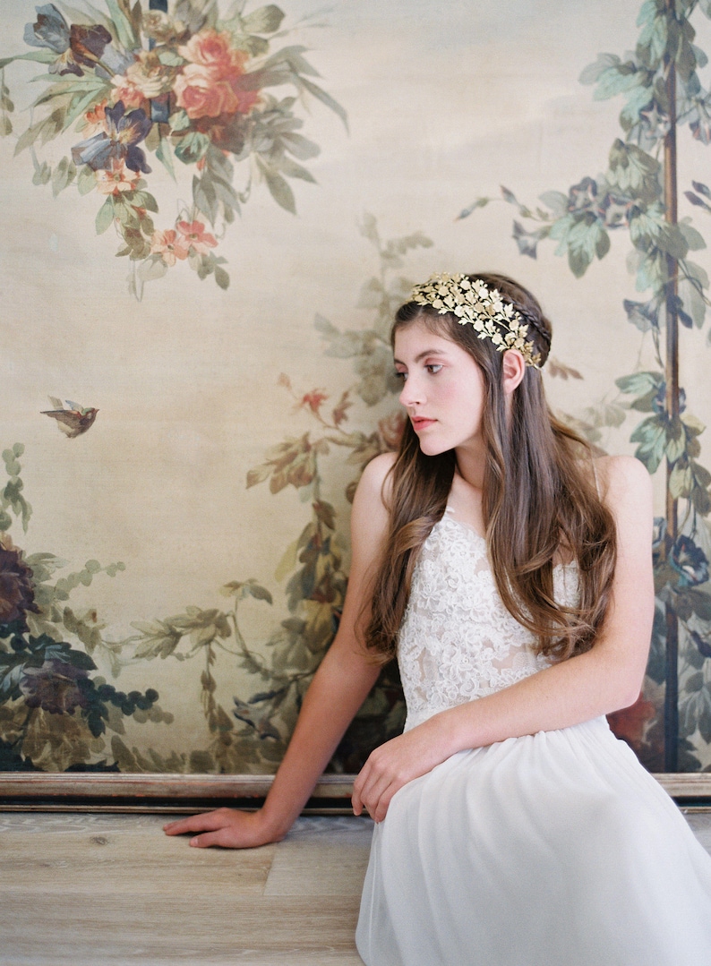 May include: A woman wearing a white lace dress and a gold leaf crown sits in front of a floral painting.