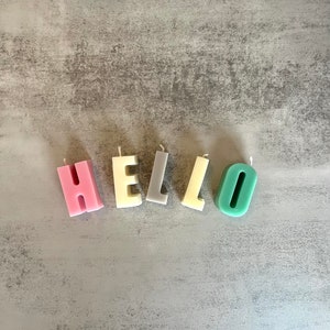 以下が含まれることがあります： 「HELLO」と綴られた文字型のキャンドル。ピンク、クリーム、グレー、イエロー、ティールなど、さまざまなパステルカラーのキャンドルが、グレーの質感のある表面に配置されています。