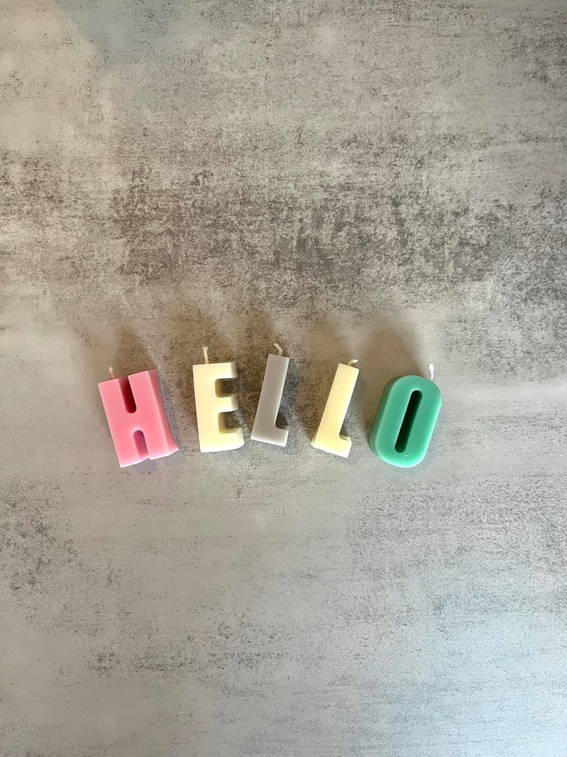 以下が含まれることがあります： 「HELLO」と綴られた文字型のキャンドル。ピンク、クリーム、グレー、イエロー、ティールなど、さまざまなパステルカラーのキャンドルが、グレーの質感のある表面に配置されています。
