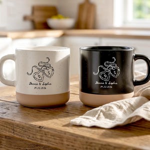 May include: Two ceramic mugs, one white and one black, each featuring a speckled finish and a tan base. The mugs display a white or black line drawing of two heart-shaped lockets with the letters D and S, the text Dennis & Sophia, and 09.12.2026.