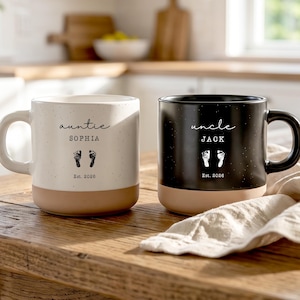 Può includere: Due tazze in ceramica, una color crema e una nera, con finitura maculata e base beige solida. La tazza color crema riporta la scritta 'auntie SOPHIA' e quella nera 'uncle JACK', entrambe con impronte di piedi di neonato e 'Est. 2026'.