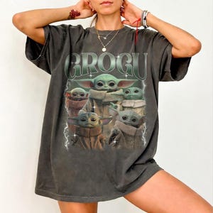 May include: Dark gray t-shirt featuring the word "GROGU" in green, with a graphic of Grogu from The Mandalorian. The graphic shows Grogu in various poses, with lightning bolts on either side. The shirt has a vintage, worn look.