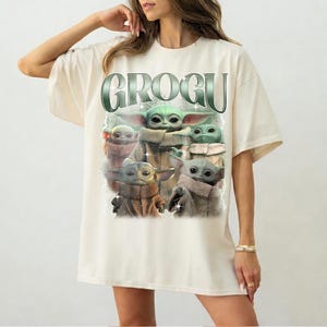 May include: Cream-colored t-shirt featuring a graphic of Grogu, with the word "GROGU" in a green, metallic font. The graphic shows multiple images of Grogu in various poses. The shirt has short sleeves and a relaxed fit.