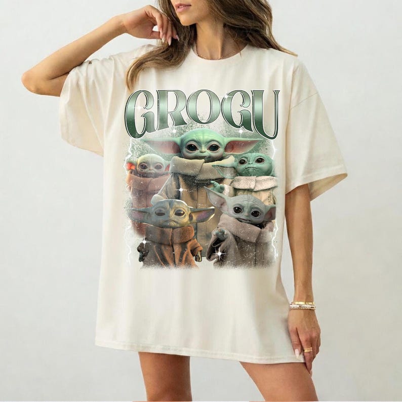 May include: Cream-colored t-shirt featuring a graphic of Grogu, with the word "GROGU" in a green, metallic font. The graphic shows multiple images of Grogu in various poses. The shirt has short sleeves and a relaxed fit.