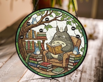 May include: Round stained glass window featuring a gray Totoro character reading a book in a cozy armchair. The scene includes stacks of books, blue creatures, and a tree with green leaves, all within a green and brown border.