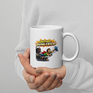 May include: White ceramic mug with a comic-style illustration of an IT professional as a superhero. The image shows a person flying with a laptop, with the text "ANOTHER DIVING CATCH!" in a yellow burst. The mug has a curved handle.