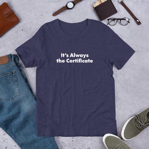 May include: Navy blue t-shirt with the white text "It's Always the Certificate". The shirt is made of a soft material and is suitable for casual wear. The shirt is displayed with jeans, shoes, and accessories.