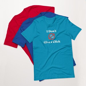 Op de afbeelding: Drie t-shirts met korte mouwen in rood, blauw en teal. Het teal shirt heeft de witte tekst "I Don't Give a Click", met een afbeelding van een cursor die is doorgestreept door een rode cirkel en lijn. De shirts zijn op elkaar gestapeld.