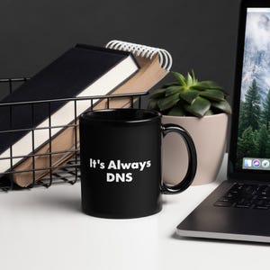 Peut inclure: Une tasse en céramique noire avec le texte "It's Always DNS" en blanc. La tasse est posée sur une surface blanche, à côté d'un ordinateur portable, d'une plante succulente dans un pot beige et d'un panier en fil de fer contenant des cahiers.