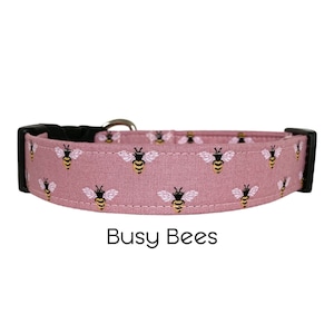 May include: A pink dog collar with a repeating pattern of black and yellow bees. The collar has a silver buckle and black D-ring. The text "Busy Bees" is printed below the collar.