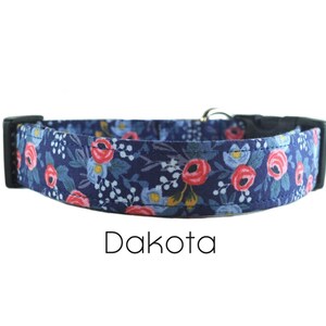 May include: A blue dog collar with a floral pattern featuring pink and white flowers. The collar has a black buckle and the text "Dakota" is printed below the collar.