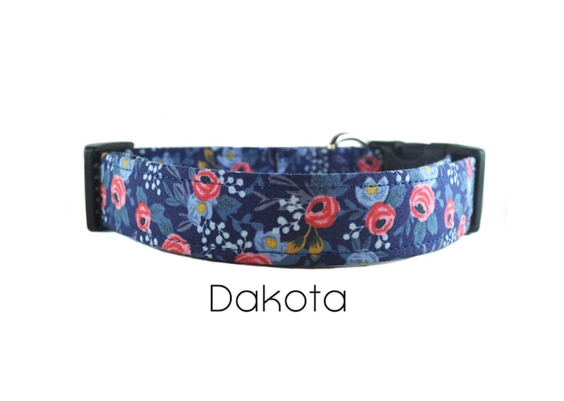 May include: A blue dog collar with a floral pattern featuring pink and white flowers. The collar has a black buckle and the text "Dakota" is printed below the collar.