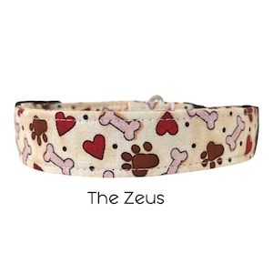 May include: A beige dog collar with a pattern of red hearts, brown paw prints, and white bones. The collar has black stitching and the text "The Zeus" is printed below the collar.