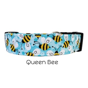 May include: A blue dog collar with a pattern of yellow and black bees on a white background. The collar has a black buckle and a silver D-ring. The text "Queen Bee" is printed below the collar.