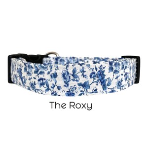 Op de afbeelding: Een hondenriem met een blauw bloemenpatroon op een witte achtergrond. De riem heeft een zwarte plastic gesp en een zilveren metalen ring. De tekst "The Roxy" is in het zwart gedrukt.