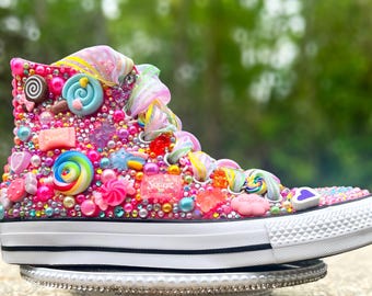 Könnte beinhalten: Ein hochgeschnittener Sneaker, der mit bunten Süßigkeiten, Perlen und Verzierungen bedeckt ist. Der Schuh hat eine weiße Gummisohle und Zehenkappe mit einer Vielzahl von Süßigkeiten in Rosa, Blau und Gelb. Der Schuh steht auf einem dekorativen Ständer.