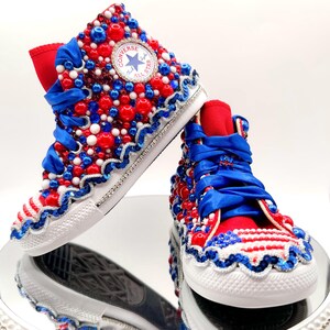 May include: A pair of high-top sneakers, embellished with red, white, and blue beads and ribbons. The sneakers feature the Converse All Star logo and a white rubber sole. The design incorporates a patriotic theme with a wavy sequin trim.