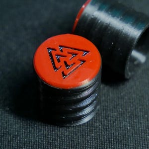 May include: Close-up of a stack of black and red cylindrical objects. The top of each cylinder is red with a black, stylized symbol. The cylinders have a ridged texture. One cylinder is separate from the stack.