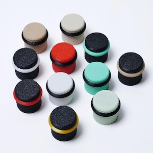 May include: A collection of colorful bottle stoppers. Each stopper has a textured, dark base and a contrasting top in various colors, including red, white, and beige. A black rubber ring encircles the top of each stopper. The stoppers are arranged on a white background.