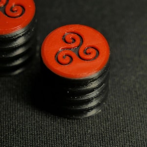 Puede incluir: Primer plano de dos objetos decorativos negros y rojos. Cada objeto es una pila de capas negras coronada por una pieza circular roja. La pieza roja presenta un diseño en espiral negro. Los objetos están sobre una superficie oscura.