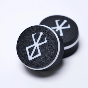 May include: A pair of circular black ear plugs featuring a textured surface and a white printed symbol resembling a stylized brand. Each plug has a distinct white band around the side, providing a high-contrast look for these round body jewelry pieces.