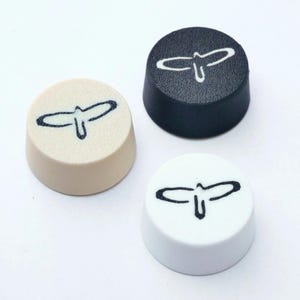 Könnte beinhalten: Drei runde, konisch geformte Knöpfe in Beige, Schwarz und Weiß. Jeder Knopf hat ein schwarzes, stilisiertes Vogel- oder Flügeldesign. Die Knöpfe sind wahrscheinlich für eine Tastatur oder ein anderes elektronisches Gerät.