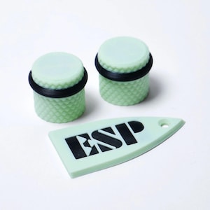 May include: Two light green cylindrical objects with a textured surface and a black ring, along with a light green tag with the letters "ESP" in black. The objects are on a white background.