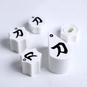 Può includere: Una collezione di gomme bianche a forma di stella e una gomma cilindrica, ciascuna con un carattere giapponese nero. Le gomme sono disposte su una superficie bianca, con un focus sul design unico e sulla forma di ogni pezzo.