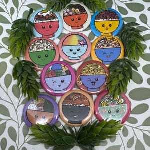 May include: A collection of colorful, round enamel pins featuring cartoon bowls of ramen and other Asian-inspired dishes. Each pin has a unique design with smiling faces and various toppings, surrounded by green foliage.