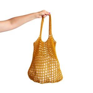 Könnte beinhalten: Senfgelbe gehäkelte Tragetasche mit langen Trägern. Das offene Webdesign zeigt ein weißes Innenfutter. Die Tasche wird von einer Hand vor weißem Hintergrund gehalten. Diese wiederverwendbare Tasche ist ideal für Lebensmittel oder zum Einkaufen.