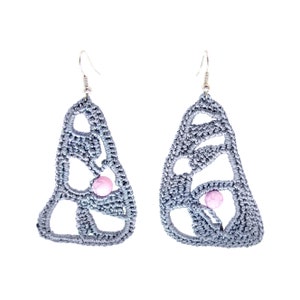 May include: A pair of handmade, gray crochet earrings with a butterfly wing design. Each earring features a small, pink bead and a silver-colored hook. The openwork design creates a delicate and airy appearance, perfect for adding a touch of bohemian flair.