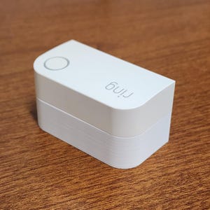 Puede incluir: Un dispositivo rectangular blanco con esquinas redondeadas, con la marca "ring" en gris. Tiene un botón circular en la parte superior y está apilado sobre capas blancas. El dispositivo está sobre una superficie de madera.