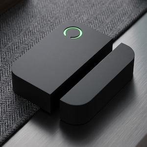 Può includere: Un sensore per la casa intelligente nero opaco con un design rettangolare arrotondato. Il sensore ha un anello verde illuminato sulla parte superiore. Il sensore è diviso in due parti, una rettangolare e una più piccola.