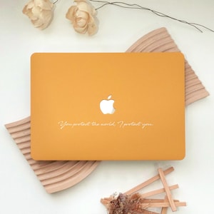 May include: Mustard yellow laptop case with a cut-out Apple logo. The case has the text "You protect the world, I protect you" in a delicate script. The case is resting on a wooden decorative element.