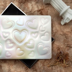May include: A white, iridescent clutch with a pattern of stitched hearts in various sizes. The clutch is open, revealing a black interior. A decorative column and dried flowers are in the background.