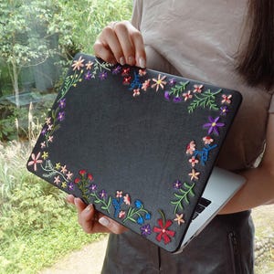 May include: A black laptop case featuring a textured surface adorned with colorful embroidered floral patterns along the edges. The embroidery includes various small flowers in shades of pink, purple, red, blue, and yellow with green stems.