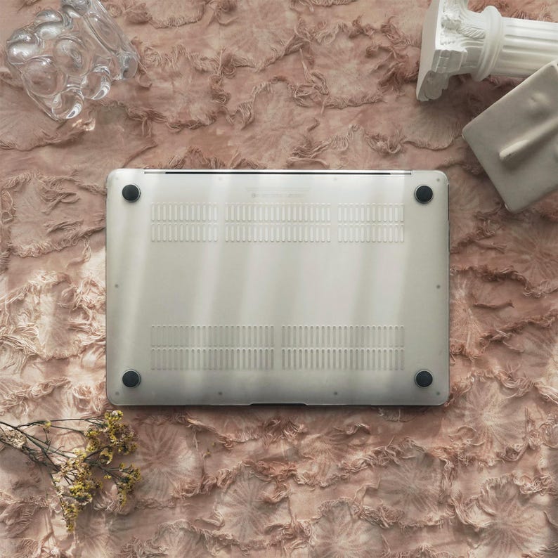 May include: A silver laptop computer with black rubber feet is centered in the image. The bottom of the laptop is visible, showing ventilation slots. Decorative items, including a glass object and a white column, are arranged around the laptop on a textured, pink surface.