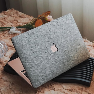 May include: A gray laptop with a textured stone-like design on the lid, featuring the Apple logo. The laptop is open, revealing a rose gold interior and keyboard. It rests on a black laptop stand, with a floral arrangement in the background.
