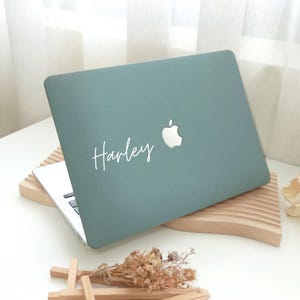 Könnte beinhalten: Ein tealfarbener Laptop mit dem Namen "Harley" in weißer Schrift. Das Apple-Logo ist sichtbar. Der Laptop steht auf einem Holzständer, die Tastatur ist sichtbar. Getrocknete Blumen und eine kleine Holzstruktur befinden sich im Vordergrund.