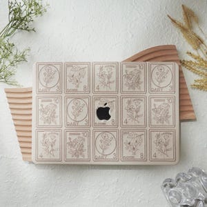 Può includere: Una custodia beige per laptop con un design a timbro floreale. La custodia presenta una griglia di timbri rettangolari con illustrazioni floreali e testo. Il logo Apple è visibile. La custodia è su un supporto di legno.