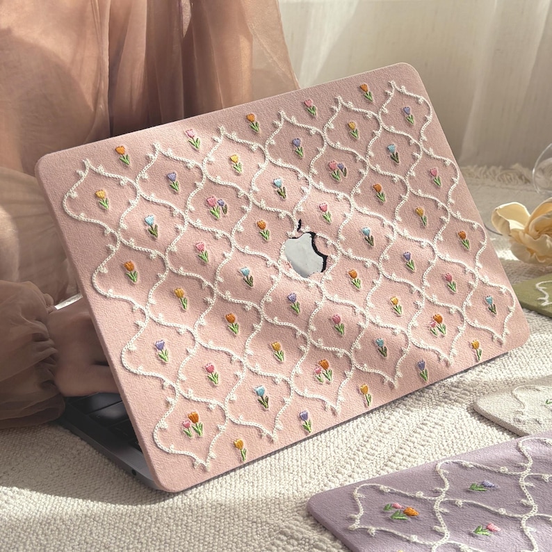May include: A pink laptop case with an embroidered floral pattern. The case features a repeating design of white, embroidered lines and small, colorful flowers. The Apple logo is visible in the center.