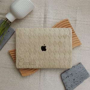 May include: A beige laptop skin with a diamond-quilted texture and a cutout for the Apple logo. The name Camille is engraved in a cursive script on the lower left corner. The skin is placed on a light-coloured fabric surface with decorative accents.