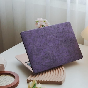 Puede incluir: Funda para portátil lila con un diseño floral en relieve. El diseño incluye flores y hojas. El portátil descansa sobre un soporte de madera curvado. Un accesorio protector.