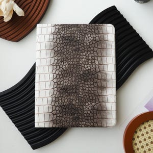 Puede incluir: Un cuaderno con un patrón de piel de cocodrilo sintética blanca y marrón. El cuaderno rectangular tiene una superficie texturizada, imitando escamas de reptil. Descansa sobre una superficie blanca, acompañado de bandejas decorativas negras y marrones. El diseño es detallado.