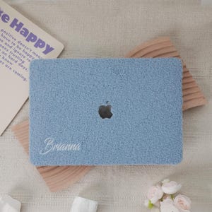 Puede incluir: Una funda para portátil azul claro con una textura difusa, con el logotipo de Apple y el nombre "Brianna" bordado en blanco. La funda está sobre un soporte de madera y una superficie beige. Se ve una tarjeta con el texto "Be Happy".